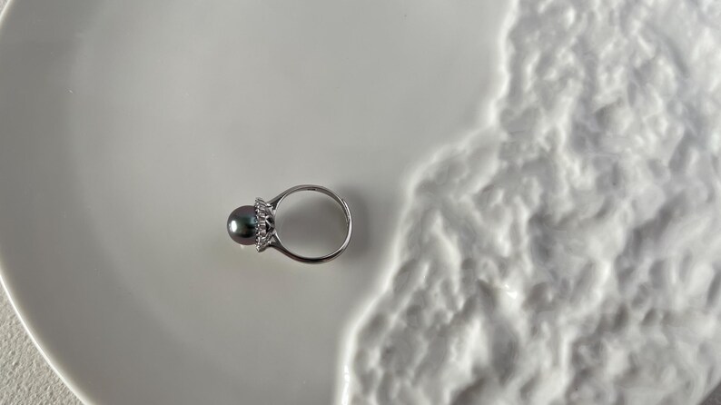 K&ouml;nnte beinhalten: Ein silberner Ring mit einem dunkelgrauen Perlmutt-Mittelstein und einem Kranz aus kleinen, klaren Edelsteinen. Der Ring liegt auf einer wei&szlig;en, strukturierten Oberfl&auml;che, die einen Kontrast erzeugt. Schmuck.