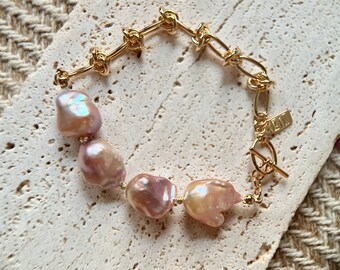 Peut inclure: Bracelet doré composé de maillons noués et ovales. Il est orné de grosses perles irrégulières rose-pêche. Une petite étiquette rectangulaire avec du texte est attachée près du fermoir.