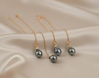 Könnte beinhalten: Goldfarbene Halsketten mit tropfenförmigen, dunkelgrauen Perlen. Die Halsketten hängen vor einem weichen, cremefarbenen Stoff. Die Perlen haben einen subtilen, irisierenden Glanz, der dem Schmuck Eleganz verleiht.