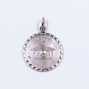 May include: A silver-colored pendant with a circular design. The pendant features an engraved image of a building with text above and below. The edges of the pendant have a decorative, scalloped pattern. The pendant has a small loop at the top for a chain.