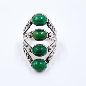 May include: A silver ring featuring four oval, vibrant green gemstones. The ring has an intricate design with decorative elements between each stone. The gemstones are set in a vertical arrangement, creating a unique and eye-catching piece of jewelry.