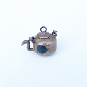 May include: A small, antique copper teapot charm with a worn finish. The teapot has a round lid and a curved spout.