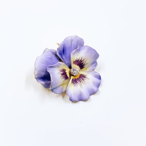 以下が含まれることがあります： パンジーの形をした花のブローチ。ラベンダーと白の色合いの花びら、黄色の中心、輝くダイヤモンドが特徴です。ブローチはおそらく金製です。