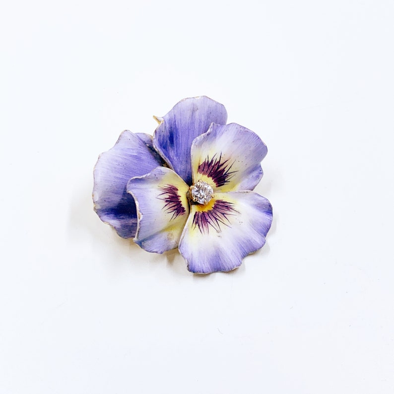 以下が含まれることがあります： パンジーの形をした花のブローチ。ラベンダーと白の色合いの花びら、黄色の中心、輝くダイヤモンドが特徴です。ブローチはおそらく金製です。