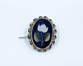 以下が含まれることがあります： 黒い背景に白い花と緑の葉がついた金色のブローチ。ブローチは楕円形で、装飾的な縁取りが施されています。