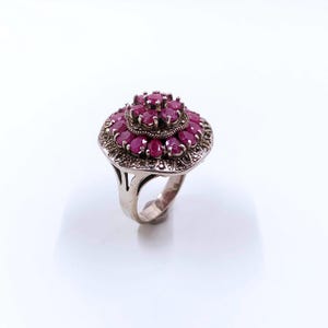 May include: A silver ring with a large, ornate setting featuring multiple red gemstones. The ring has a delicate, openwork band.