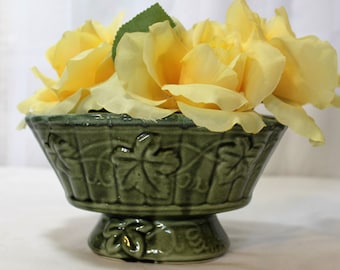 May include: A green ceramic planter with a vine design, holding a bouquet of yellow roses.