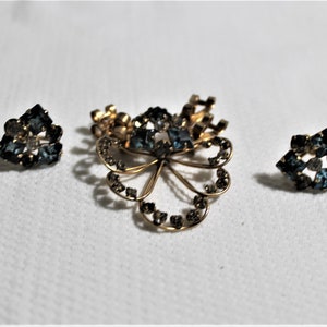May include: A set of vintage jewelry featuring a gold-tone brooch with a floral design and blue rhinestones, and two matching earrings with blue rhinestones.
