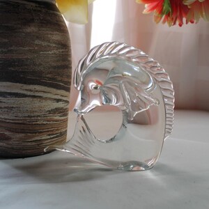 May include: A clear glass sculpture of a fish with a curved tail and a detailed head. The sculpture is sitting on a white surface.