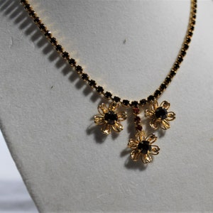 Puede incluir: Un collar de oro con una cadena de pedrer&iacute;a negra y tres colgantes de flores. Las flores tienen pedrer&iacute;a negra en el centro y detalles en metal dorado.