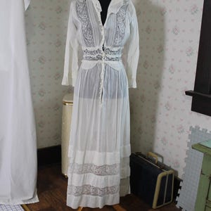 May include: A white vintage dress on a black mannequin. The dress features long sleeves, a collared neckline, and intricate lace detailing on the bodice and skirt. The dress is floor-length and has a button-down front.