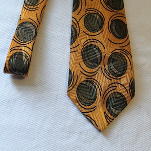 May include: A gold tie with a black and brown geometric pattern. The pattern features circles with a swirling design.