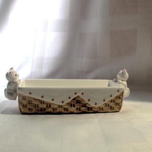 May include: A rectangular ceramic basket-style container with a white interior and a woven brown exterior. Two small white cat figurines are attached to the sides. The top edge has a white trim with red cherry accents. A decorative piece.