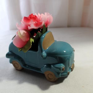 May include: A teal blue ceramic truck planter with pink and white flowers. The truck has a brown windshield and a detailed front grill.