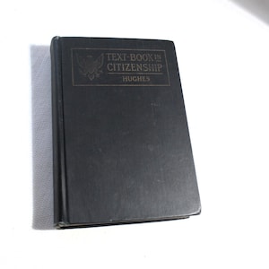 May include: A black textbook titled "TEXT-BOOK IN CITIZENSHIP" with the author's name, Hughes, printed in gold lettering. The book has a dark cover and appears to be in good condition. The book is on a white surface.