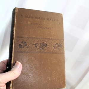 May include: A vintage brown book with the title "MIRIAM COLES HARRIS" and "AUTHOR OF 'RUTLEDGE'" printed on the cover. The cover features a decorative floral design. The book is held in a hand.