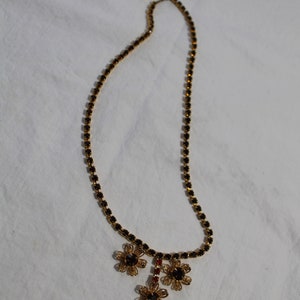 Puede incluir: Un collar de oro con una cadena de peque&ntilde;os cristales negros y un colgante de tres abalorios de oro en forma de flor con cristales negros.