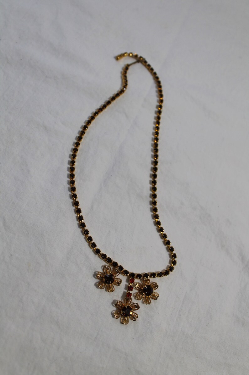 Puede incluir: Un collar de oro con una cadena de peque&ntilde;os cristales negros y un colgante de tres abalorios de oro en forma de flor con cristales negros.