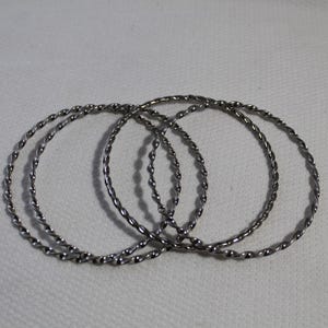 May include: Three silver-toned metal bangles, each featuring a continuous twisted rope texture. The circular bracelets are arranged in an overlapping formation, showcasing the reflective, metallic surface and the uniform, repeating spiral pattern.