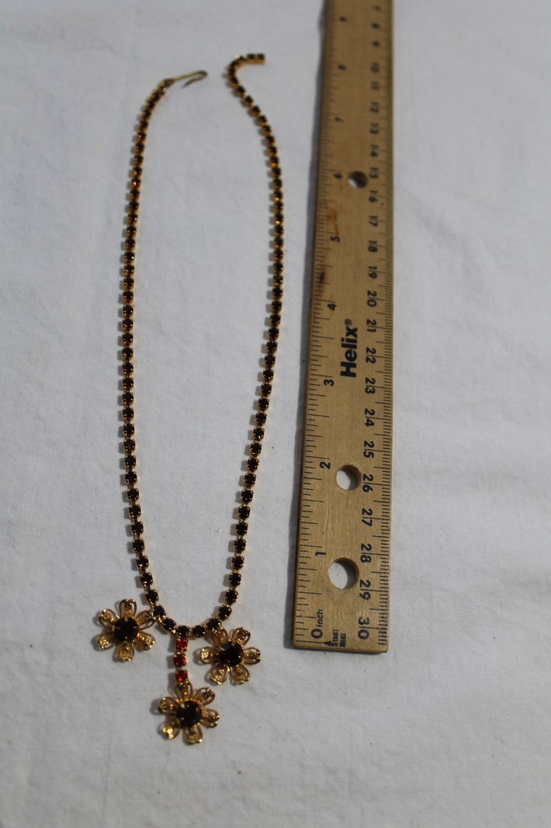 Puede incluir: Un collar de oro con una cadena de peque&ntilde;os diamantes de imitaci&oacute;n negros y un colgante de tres flores doradas con diamantes de imitaci&oacute;n negros. El collar mide 45,7 cm de largo.