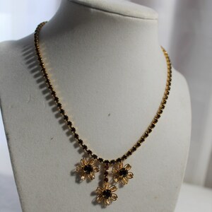 Puede incluir: Un collar de oro con un colgante de flor de pedrer&iacute;a negra y roja. El collar est&aacute; en un busto de maniqu&iacute; blanco.