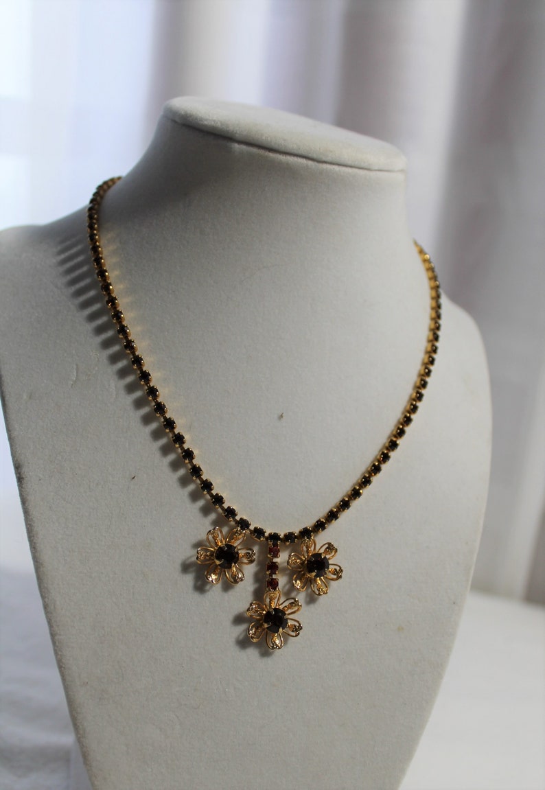 Puede incluir: Un collar de oro con un colgante de flor de pedrer&iacute;a negra y roja. El collar est&aacute; en un busto de maniqu&iacute; blanco.