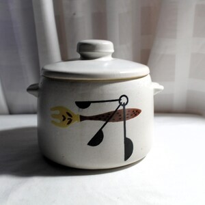 May include: A cream-colored ceramic casserole dish with a lid, featuring a whimsical design of a fork, spoon, and ladle in yellow, brown, and black. The dish has two small handles and is suitable for kitchen use.