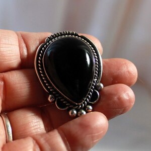 May include: A silver ring with a large, teardrop-shaped black stone. The stone is set in a decorative silver frame with a twisted rope design and small silver beads. The ring is held in a hand.