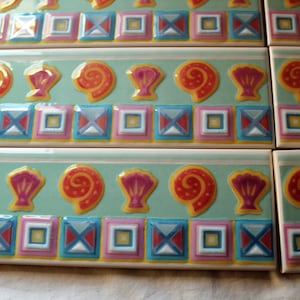 May include: A set of four ceramic tile trim pieces with a repeating pattern of seashells, swirls, and geometric shapes in shades of pink, yellow, blue, red, and green on a light blue background.
