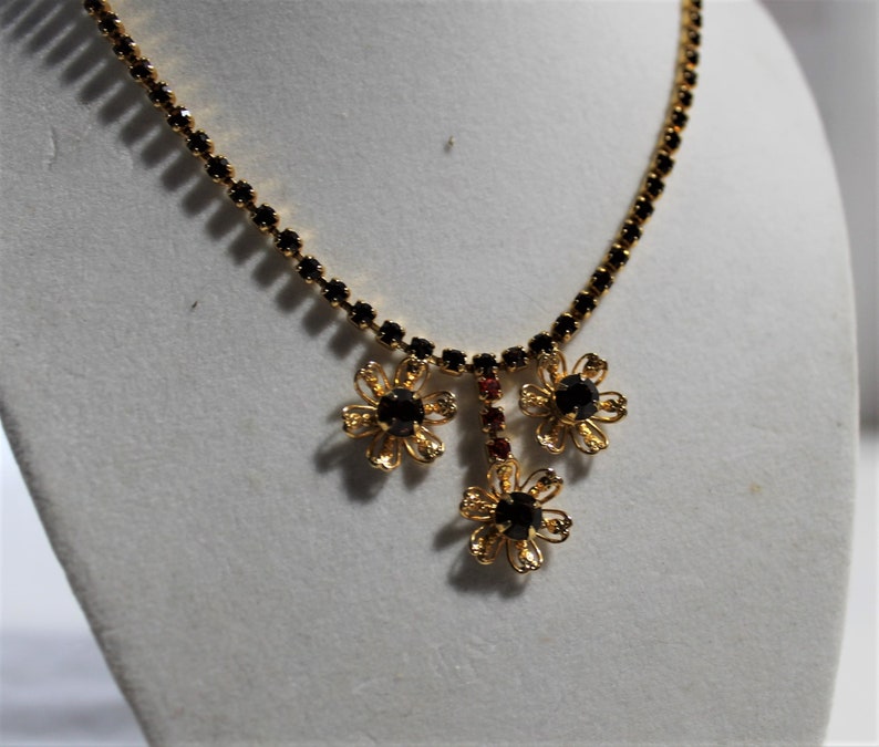 Puede incluir: Un collar de oro con una cadena de pedrer&iacute;a negra y tres colgantes de flores. Las flores tienen pedrer&iacute;a negra en el centro y detalles en metal dorado.