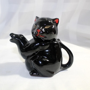 May include: Black ceramic teapot shaped like a cat with red ears and a spout. The teapot is sitting on a white surface.