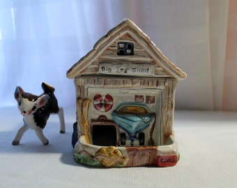 May include: A ceramic toy shed with a brown roof and a blue door. The shed has a sign that says "Big Toy Shed" and a small deer figurine is in front of it.