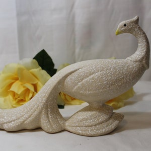 May include: A white ceramic peacock figurine with a textured finish. The peacock is standing with its tail feathers spread out behind it.