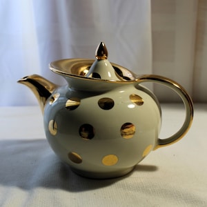 May include: A white teapot with gold polka dots and a gold lid. The teapot has a round body and a curved handle.