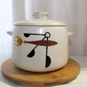 May include: White ceramic crock pot with a lid and a brown, black, and yellow design of kitchen utensils. The crock pot is sitting on a wooden base.