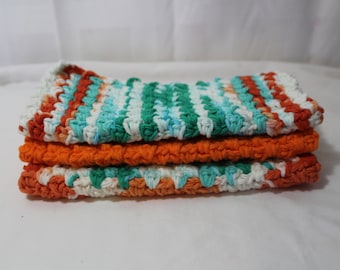 May include: A stack of four crocheted dishcloths in orange, white, and teal colors. The dishcloths are made with a textured stitch pattern.