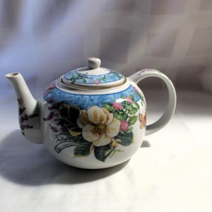 May include: A white porcelain teapot with a floral design featuring pink, yellow, and blue flowers. The teapot has a blue and white patterned lid and a handle.