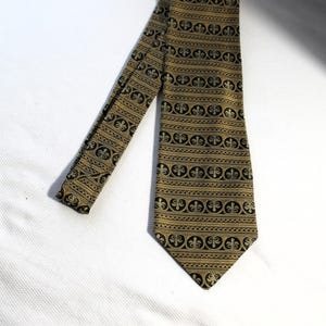 May include: A gold and black patterned tie. The tie features a repeating pattern of black fleur-de-lis shapes and horizontal stripes. The tie is made of a shiny material and is displayed on a white surface.