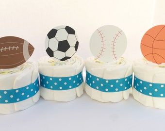 May include: Four diaper cakes decorated with blue and white polka dot ribbon and topped with sports-themed cutouts: a football, soccer ball, baseball, and basketball.