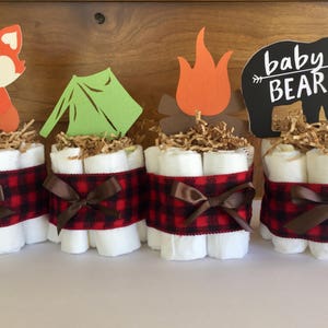 May include: Four diaper cakes decorated with a woodland theme. Each cake is wrapped in red and black buffalo plaid ribbon and topped with a different woodland animal cutout. The cutouts include a fox, a tent, a campfire, and a bear with the text "baby bear".