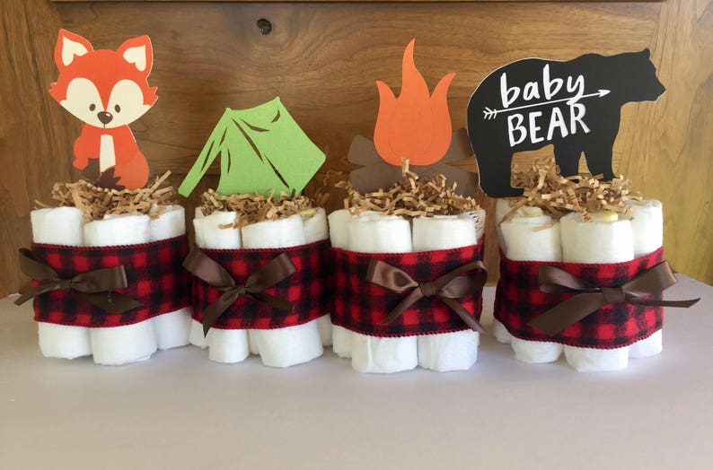 May include: Four diaper cakes decorated with a woodland theme. Each cake is wrapped in red and black buffalo plaid ribbon and topped with a different woodland animal cutout. The cutouts include a fox, a tent, a campfire, and a bear with the text "baby bear".