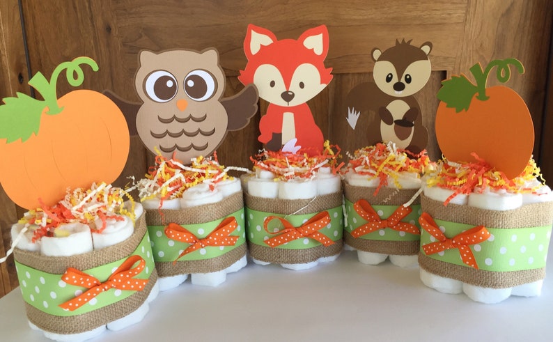 May include: Four diaper cakes decorated with burlap ribbon, green and orange polka dot ribbon, and orange bows. Each cake is topped with a different woodland animal cutout: an owl, a fox, a squirrel, and a pumpkin.