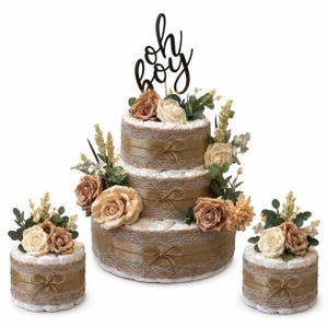 May include: A three-tier diaper cake and two smaller single-tier diaper cakes decorated with burlap, lace ribbon, gold satin bows, and artificial cream and tan roses with green foliage. A black script cake topper reads oh boy.