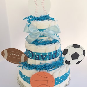 May include: A blue and white diaper cake decorated with sports-themed cutouts. The cake is topped with a white baseball, and the bottom tier has a basketball, a football, and a soccer ball. The cake is decorated with a blue ribbon and the name "David" in blue letters.