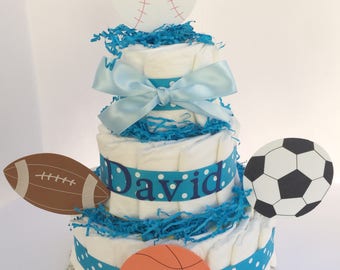 May include: A blue and white diaper cake decorated with sports-themed cutouts. The cake is topped with a white baseball, and the bottom tier has a basketball, a football, and a soccer ball. The cake is decorated with a blue ribbon and the name "David" in blue letters.
