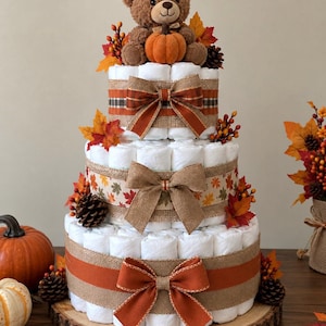 May include: A three-tiered diaper cake constructed from rolled white diapers, decorated with burlap and orange ribbons, bows, artificial autumn leaves, pinecones, and berries. A plush brown teddy bear holding a small orange pumpkin sits on top.