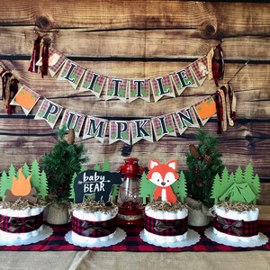 May include: A woodland themed baby shower centerpiece with three diaper cakes decorated with plaid ribbon, faux greenery, and woodland animal cutouts. The cakes are topped with a campfire, a fox, and a tent. A banner reads "Little Pumpkin" with plaid accents.