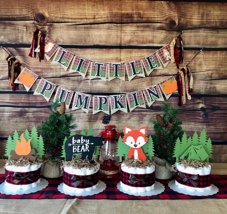 May include: A woodland themed baby shower centerpiece with three diaper cakes decorated with plaid ribbon, faux greenery, and woodland animal cutouts. The cakes are topped with a campfire, a fox, and a tent. A banner reads "Little Pumpkin" with plaid accents.