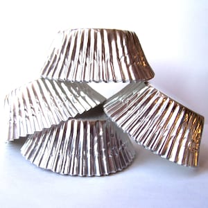 May include: A stack of four silver cupcake liners, each with a fluted design. The liners are made of aluminum foil and are designed to be used in a muffin tin.