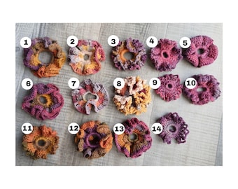 May include: A collection of fourteen handmade crochet scrunchies in various colors, including orange, purple, pink, and beige. Each scrunchie has a unique textured design and is numbered for easy selection. The scrunchies are displayed on a light gray wooden surface.